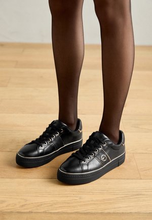 Benen met doorschijnende zwarte panty's en zwarte leren sneakers met gouden veteroogjes en een klein logo op een lichte houten vloer.