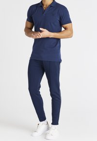 Mies, který má na sobě tmavě modré polo tričko a odpovídající slim-fit teplákové kalhoty s bílými teniskami na jednolitém pozadí.