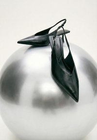 Mustat teräväkärkiset nilkkarihvelit lepäävät suurella heijastavalla hopeisella pallolla yksinkertaisella taustalla.