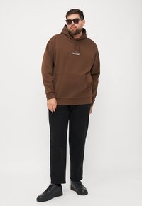 Hanorac maro cu buzunar tip kangaroo și logo brodat. Asociat cu pantaloni negri și adidași negri. Ochelarii de soare completează look-ul.