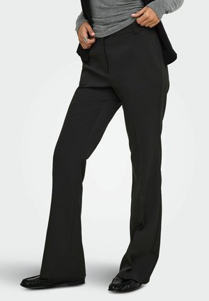 Femme portant un pantalon noir droit, un haut gris à manches longues, des ballerines noires, les mains ajustant la zone de la taille.