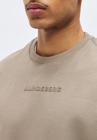 Κοντινή εικόνα ενός ατόμου που φοράει μπεζ φούτερ με στρογγυλή λαιμόκοψη και ανάγλυφο κείμενο "J.LINDEBERG" στο στήθος.