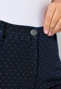 Tmavě modré džíny s přiléhavým pasem, zdobené stříbrným knoflíkem a bílým tečkovaným vzorem. Detailní záběr zaměřený na texturu a designové detaily.