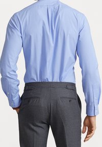 Man som bär en ljusblå skjorta instoppad i mörkgrå byxor, står med händerna avslappnat vid sidorna, vänd bort från betraktaren.