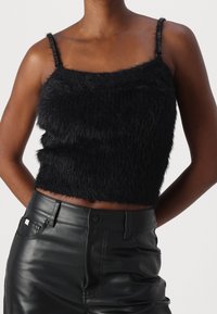 Persona che indossa un crop top nero senza maniche e peloso con sottili spalline e pantaloni in pelle nera a vita alta, con le braccia posizionate dietro la schiena.