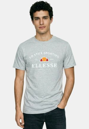 Jeune homme aux cheveux bouclés portant un t-shirt Ellesse gris clair avec logo et texte, jean foncé, posant avec une main dans la poche.