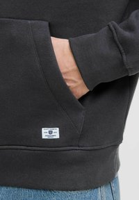 Felpa grigio scuro con tasca a marsupio, polsini e orlo a coste. Presenta un'etichetta con logo bianca con testo blu vicino alla tasca.
