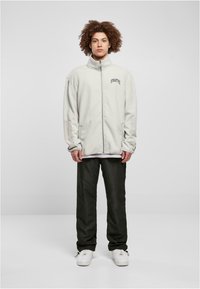 Vaaleanharmaa fleece-zip-up-takki, jossa on korkea kaulus ja brodeerattu teksti, yhdistetty tummiin housuihin ja valkoisiin lenkkareihin.