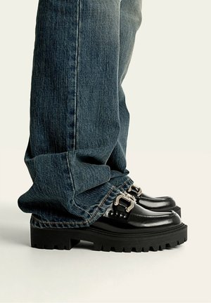 Zwarte chunky plateau loafers met zilveren gespen, gedragen met lange, losse donkerblauwe spijkerbroek die de enkels bedekt.