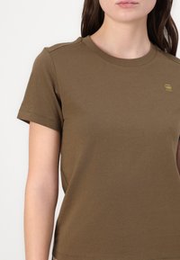 T-shirt brun à manches courtes en coton avec un col rond. Présente un petit logo doré clair sur la poitrine. Texture lisse.