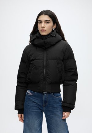 Donna con lunghi capelli scuri che indossa orecchini a cerchio d'argento, un piumino nero con collo alto e jeans blu, in piedi davanti a uno sfondo semplice.