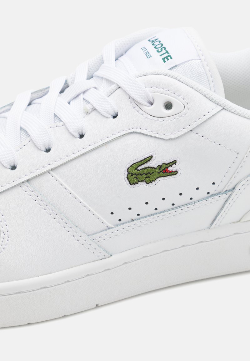 White leather sneaker with textured surface, flat laces, perforations, and green crocodile logo on the side. Rubber sole, minimal design.