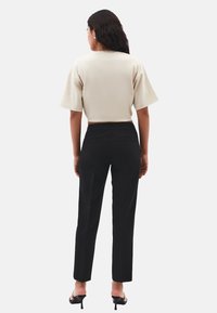 Mujer con pelo largo y oscuro, vestida con una camiseta corta beige, pantalones negros ajustados y sandalias de tacón negras, de pie con la espalda hacia delante.