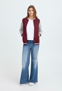 Burgunder- och grå bomberjacka med ribbad krage och mudd, kombinerad med ljusblå flare-jeans och vita sneakers. Enkel, avslappnad design.