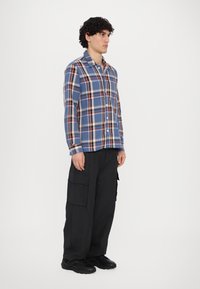 Knappete flanellskjorte i blått med rød og gul rutete mønster. Lange ermer, krage og avslappet passform. Kombinert med svarte cargo-bukser og joggesko.