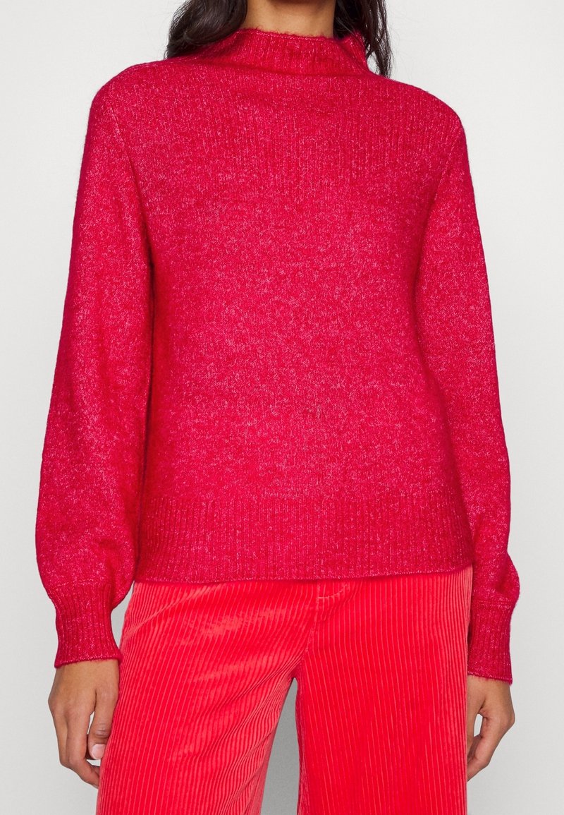 Maglione rosso a maglia con colletto alto e polsini a costine, caratterizzato da una texture morbida e una leggera lucentezza, abbinato a pantaloni in velluto a coste rossi.