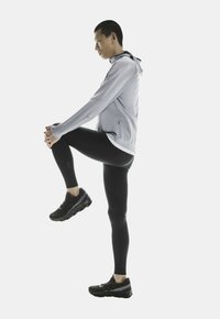 Veste de sport à capuche gris clair et leggings noirs, avec un tissu texturé et des chaussures de sport. La silhouette démontre un étirement.