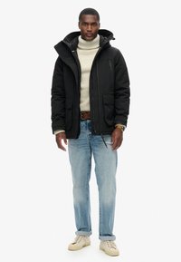 Chaqueta de plumas negra con capucha, cierre de cremallera y bolsillos frontales. Llevada sobre un suéter de cuello alto beige y combinada con jeans de un azul claro y zapatillas crema.