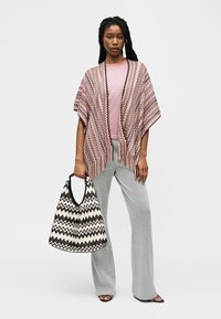 Femme portant un châle à motif zigzag rose, un haut rose, un pantalon gris, tenant un grand sac à motif zigzag noir et blanc.