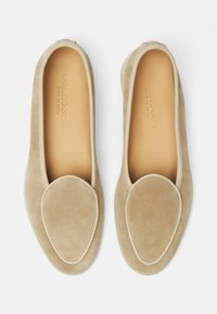 Zapatos mocasines de ante beige con punta redondeada, textura suave, acento de costura blanca y suela flexible. El interior presenta una marca de logo.