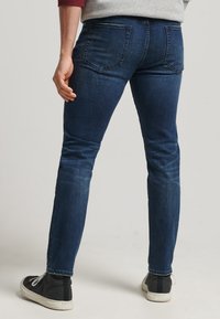 Vaqueros de mezclilla azul oscuro con un corte slim, altura media y bolsillos traseros. Llevados con zapatillas negras de caña alta y detalles de costura expuesta.