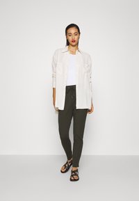 Lichtgewicht, beige lange mouw shirt over een witte tanktop; donkergroene broek met een trekkoord; zwarte sandalen met stippenaccenten.