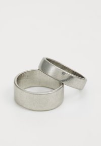 Två silvermetallringar av varierande bredd. Den större har en matt yta, medan den mindre är polerad med lätt yta och imperfektioner.