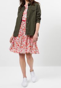 Olivengrøn arbejdstjakke over en blomstrete pink kjole med røde detaljer, der har plisseringer. Parret med hvide sneakers for et afslappet look.