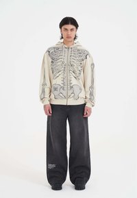 Felpa crema con grafica di uno scheletro, chiusura a zip frontale e cappuccio con cordino; abbinata a pantaloni in denim scuri e larghi e calzature nere.