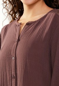 Camicia button-up marrone, con texture, caratterizzata da un colletto a V, cinque bottoni sulla parte anteriore e un leggero motivo stropicciato.