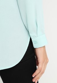 Blusa de mangas largas en aqua claro con una textura suave, cierre de botones en los puños y un dobladillo redondeado, combinada con pantalones ajustados negros.