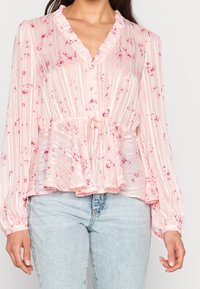Blusa rosa chiaro con scollo arricciato, righe verticali e motivi floreali. Presenta maniche lunghe e una vita arricciata con coulisse.
