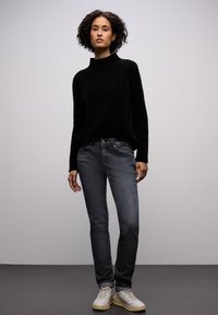 Černý svetr s vysokým límcem ve spojení s tmavě šedými džínami a bílými teniskami. Oblečení má volný střih a hladkou texturu.