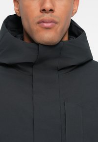 Černá kapucňová bunda s hladkým, matným povrchem, vybavená zapínáním na zip a boční kapsou. Minimalistický design zdůrazňuje funkčnost.