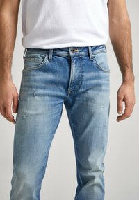 Hombre vestido con jeans azules desgastados y una camiseta blanca metida por dentro, de pie con los brazos relajados a los lados.