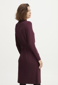 Bordowa sukienka z długim rękawem, dopasowanym paskiem w talii, miękką teksturą i długością do kolan. Prosty design i minimalne akcenty.