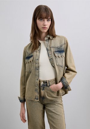 Chaqueta vaquera en colores azul descolorido y crema, con dos bolsillos en el pecho y costuras con patrón de ondas. Combinada con una camiseta ajustada crema y vaqueros.