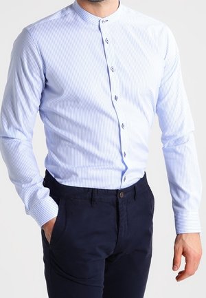 Camicia azzurro chiaro a maniche lunghe con righe verticali bianche, colletto a fascia e sei piccoli bottoni. Indossata con pantaloni scuri.