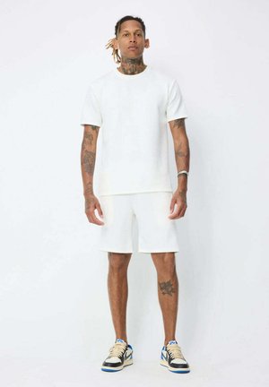 Homme avec des tatouages portant un t-shirt blanc et un short, debout devant un fond blanc uni avec des baskets bleues et blanches.