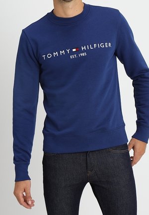 Mężczyzna noszący niebieską bluzę Tommy Hilfiger z logo i napisem „EST. 1985”, zestawioną z ciemnoniebieskimi dżinsami.