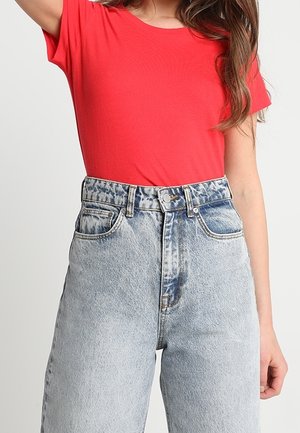 Kobieta ubrana w jasnoczerwoną koszulkę z krótkim rękawem, wpuszczoną w jeansy z wysokim stanem w jasnym odcieniu z efektem sprania.