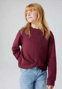Felpa bordeaux con design frontale attorcigliato, maniche lunghe, polsini a coste e texture liscia, abbinata a jeans azzurri chiari.