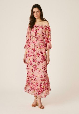 Mujer con un vestido rosa de flores con los hombros descubiertos y cinturón, de pie con expresión neutra y sandalias beige de tacón, sobre un fondo liso.