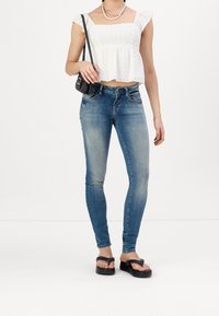 Bílý top s výšivkou a volánovými rukávy, zkombinovaný s úzkými modrými džíny. Černá crossbody taška a ploché platformové sandály dotvářejí outfit.