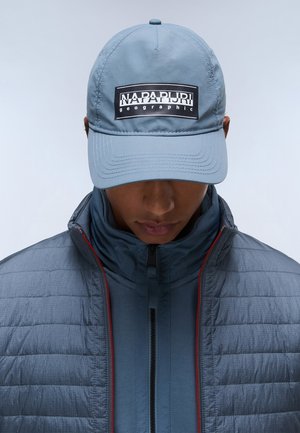 Grijze pet met een zwart logo patch, gecombineerd met een grijze pufferjack met een gladde textuur en rode accenten. Close-up van de kleding.