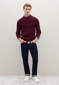 Maglione bordeaux a maglia grossa, jeans blu scuro e scarpe da ginnastica bianche. Il maglione ha un collo alto; i jeans hanno risvolti e cuciture a contrasto.
