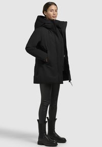 Zwarte parka met een oversized capuchon, ritsluiting en zijzakken, gecombineerd met zwarte legging en enkellaarsjes. Soepele textuur, modern ontwerp.