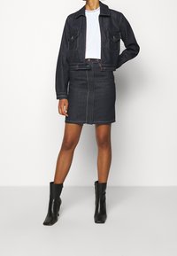 Mörk denimkortsjacka med två bröstfickor och kontrastsömmar, tillsammans med en mörk denimpennkjol och svarta ankelskor.