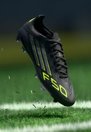 Chaussure de football noire avec texte et bandes jaune néon « F50 », flottant au-dessus de l’herbe sur un terrain de football avec des lignes blanches de délimitation.