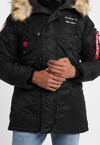 Parka noire isolante avec capuche en fausse fourrure, dotée de boutons et de patchs, avec un accent rouge sur la manche gauche. Tissu texturé avec une construction robuste.